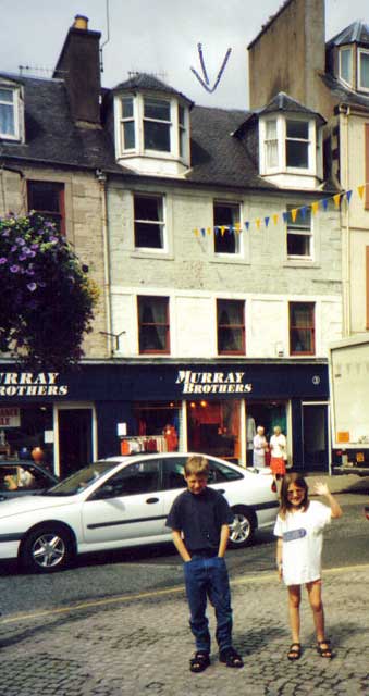 Two great-great-great grandchildren of George Oman, standing in front of 3 High Street, Hawick, the premises used by George Oman as a tobacconist's shop in 1875.